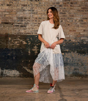 A woman stands indoors against a brick wall, smiling as she holds her skirt, dressed in a beige t-shirt, sheer lace skirt layered over shorts, and statement sneakers—the Breath sneakers by Bed|Stü.