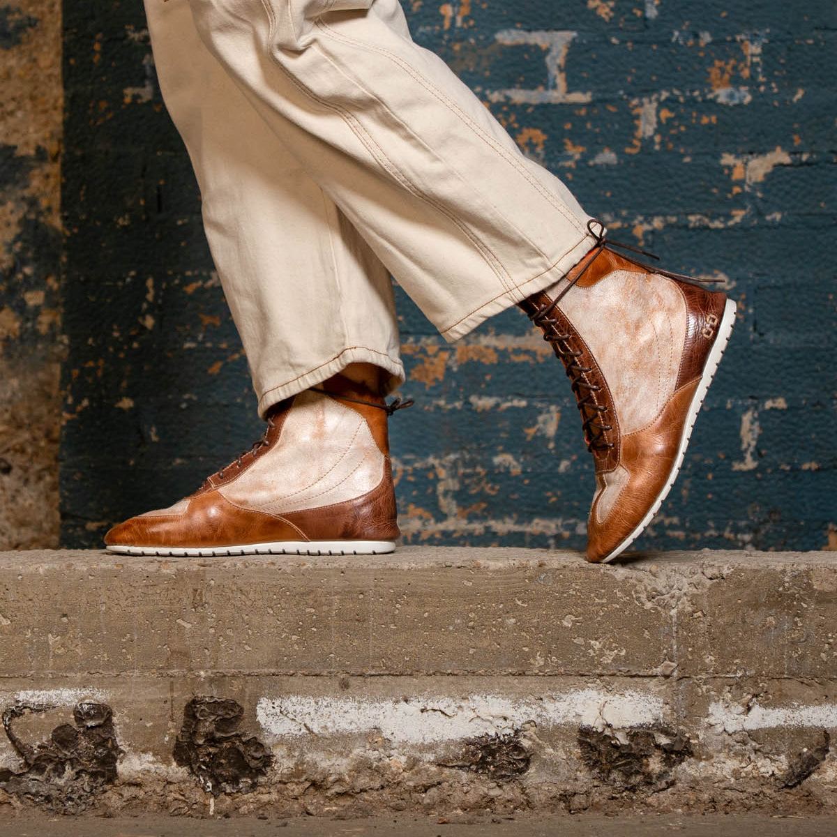 Wearing Bed|Stü's Breath statement sneakers, a person stands on a concrete step in tan pants, framed by a textured blue brick wall.