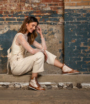 A woman sits on a concrete step against a worn brick wall, smiling with her head tilted and hand in her hair, styled in cream overalls and a sheer lace top, and wearing the Bed|Stü Breath sneakers—statement footwear featuring retro court styling and multi-tone leather details.