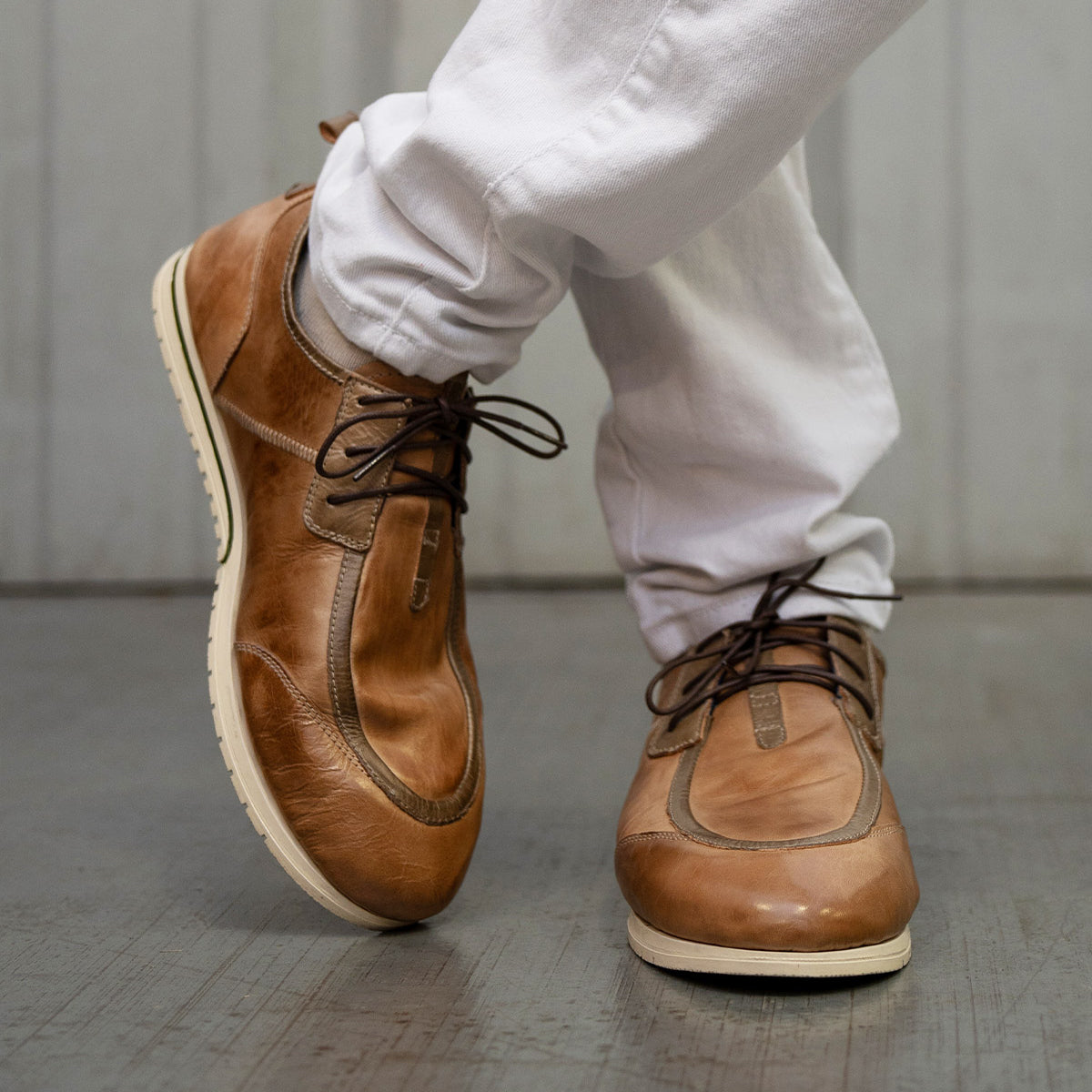 A person wearing white pants and refined brown leather Bed|Stü Britches chukka-style lace-up shoes stands on a wooden floor with one foot crossed over the other.