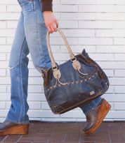 A person wearing blue jeans and brown wedge shoes carries the Bruna bag by Bed|Stü—a large, two-toned leather bag with padded straps—in front of a white brick wall.