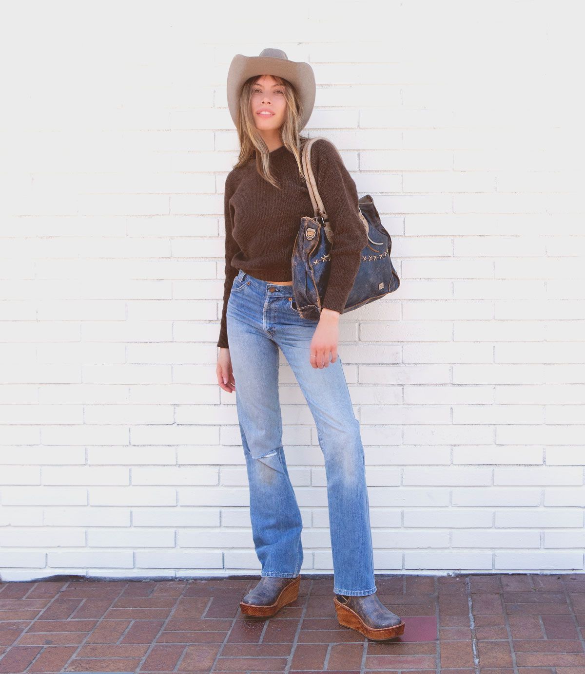 A person in a cowboy hat, brown sweater, blue jeans, and platform shoes stands against a white brick wall, holding the Bruna crossbody shoulder bag by Bed|Stü, featuring padded straps.