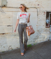 A woman stands on the sidewalk by a white brick wall, wearing sunglasses, a "Crema" graphic t-shirt, striped pants, and holding the Bed|Stü Bruna—a chic two-toned leather bag with padded straps.