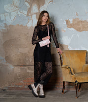 A woman wearing a sheer black lace dress and platform boots stands beside a worn yellow armchair against a distressed wall, holding the Cadence crossbody clutch by Bed|Stü in pink.