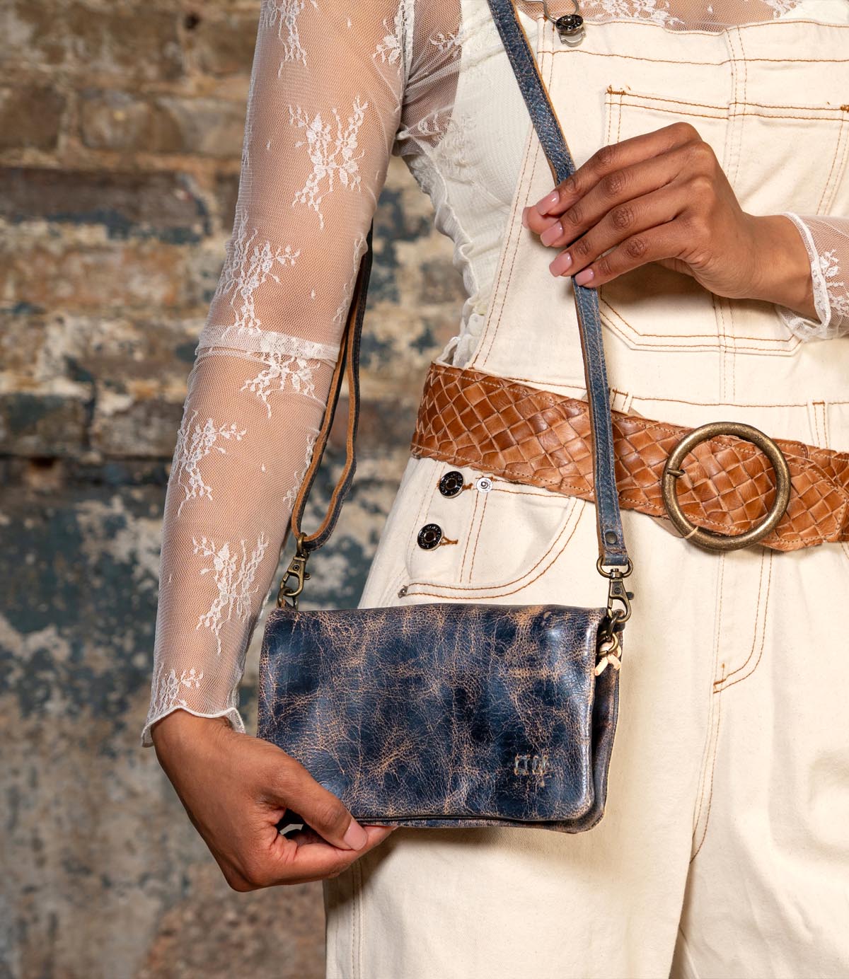 A person stands against a textured brick wall wearing a white lace top and cream overalls with a brown woven belt, holding the Bed|Stü Cadence vegetable-tanned leather crossbody clutch in distressed blue.