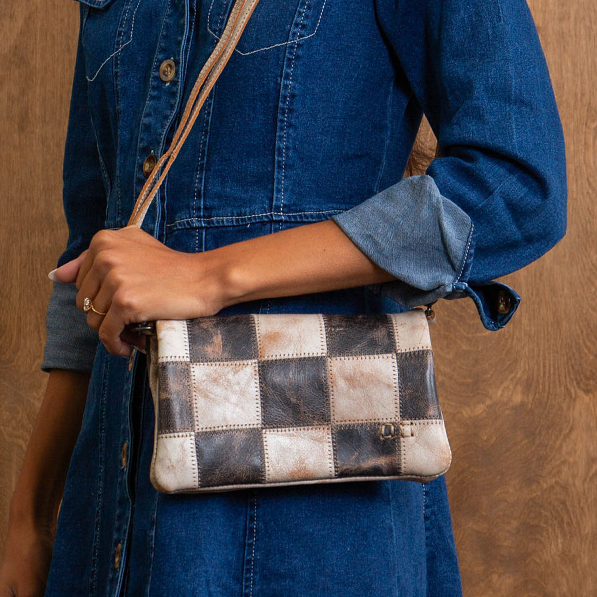 A person wearing a denim dress holds the Bed|Stü Cadence PW crossbody bag—crafted from vegetable-tanned leather with brown and beige checkered squares—against a wooden background.