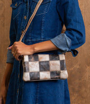 A person wearing a denim dress holds the Bed|Stü Cadence PW crossbody bag—crafted from vegetable-tanned leather with brown and beige checkered squares—against a wooden background.
