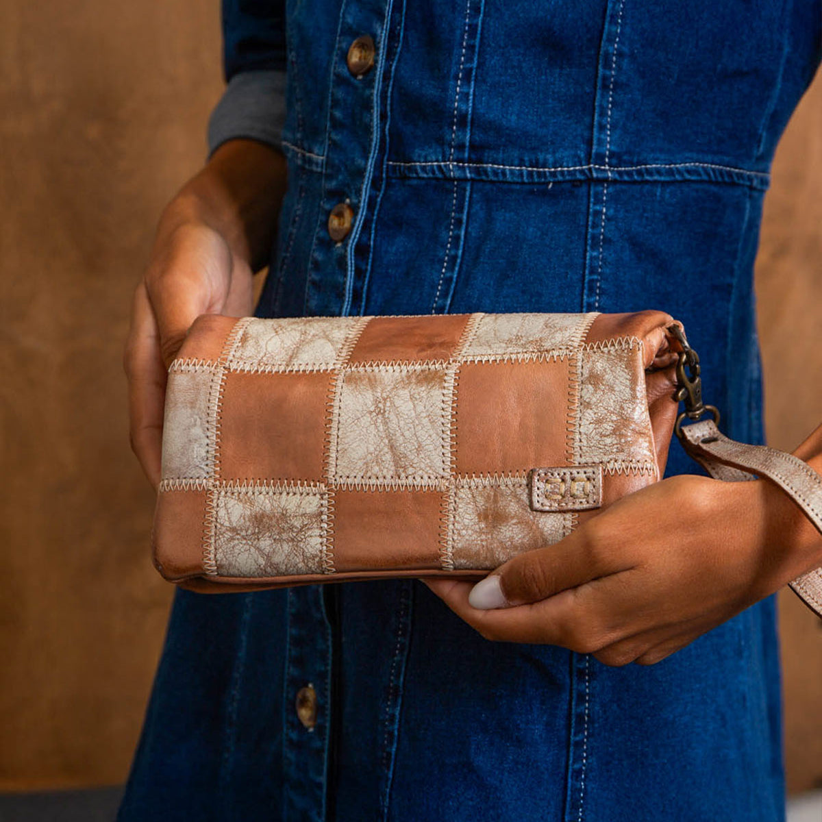 A person in a blue denim dress holds the Cadence PW patchwork clutch bag by Bed|Stü, featuring a checkered pattern crafted from vegetable-tanned leather in shades of tan and beige.