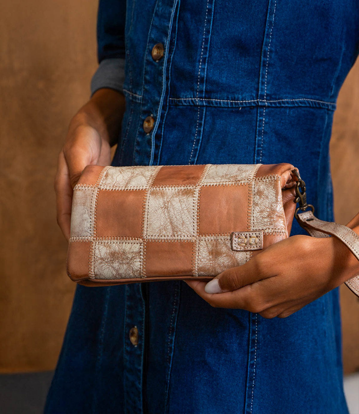 A person in a blue denim dress holds the Cadence PW patchwork clutch bag by Bed|Stü, featuring a checkered pattern crafted from vegetable-tanned leather in shades of tan and beige.