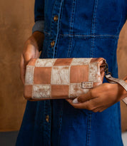 A person in a blue denim dress holds the Cadence PW patchwork clutch bag by Bed|Stü, featuring a checkered pattern crafted from vegetable-tanned leather in shades of tan and beige.