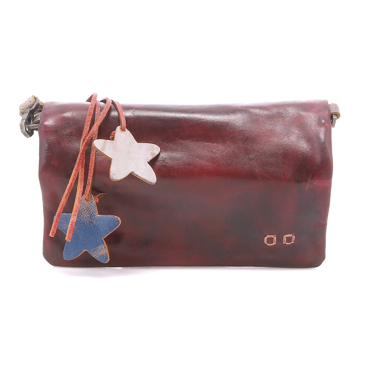 The Cadence by Bed|Stü is a brown shoulder bag crafted from supple vegetable-tanned leather, featuring two star-shaped charms—one white and one blue—attached by leather straps. This versatile accessory can also be styled as a crossbody clutch for added convenience.