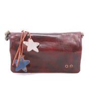 The Cadence by Bed|Stü is a brown shoulder bag crafted from supple vegetable-tanned leather, featuring two star-shaped charms—one white and one blue—attached by leather straps. This versatile accessory can also be styled as a crossbody clutch for added convenience.