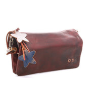 Introducing Cadence by Bed|Stü: a brown crossbody clutch bag, exquisitely crafted from vegetable-tanned leather and adorned with two star-shaped charms—one white and one blue—gracefully attached to the side.