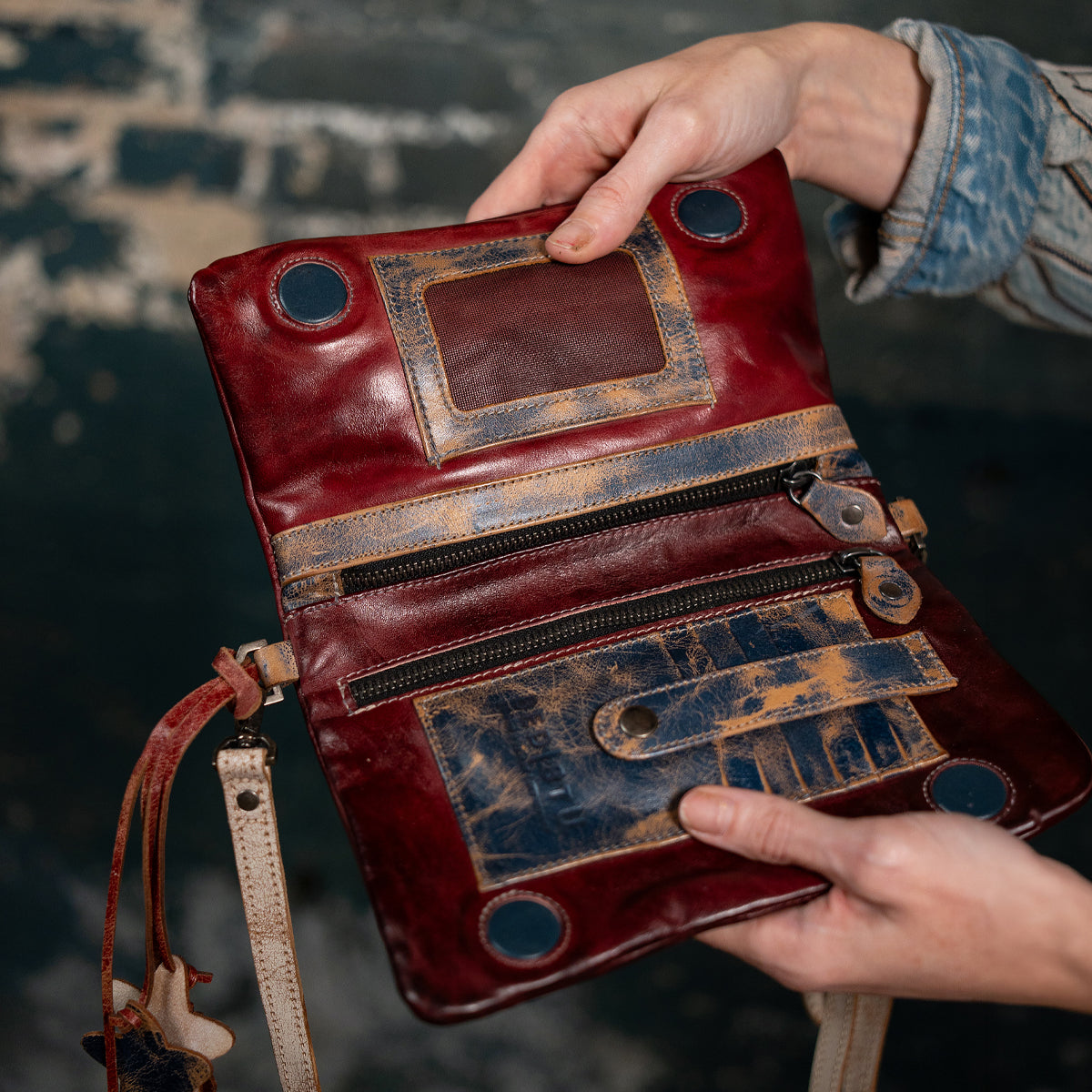 A person holding the Bed|Stü Cadence, an open vintage-style purse crafted from vegetable-tanned leather, showcasing visible zippers, roomy pockets, and distressed brown and red accents against a softly blurred dark background.