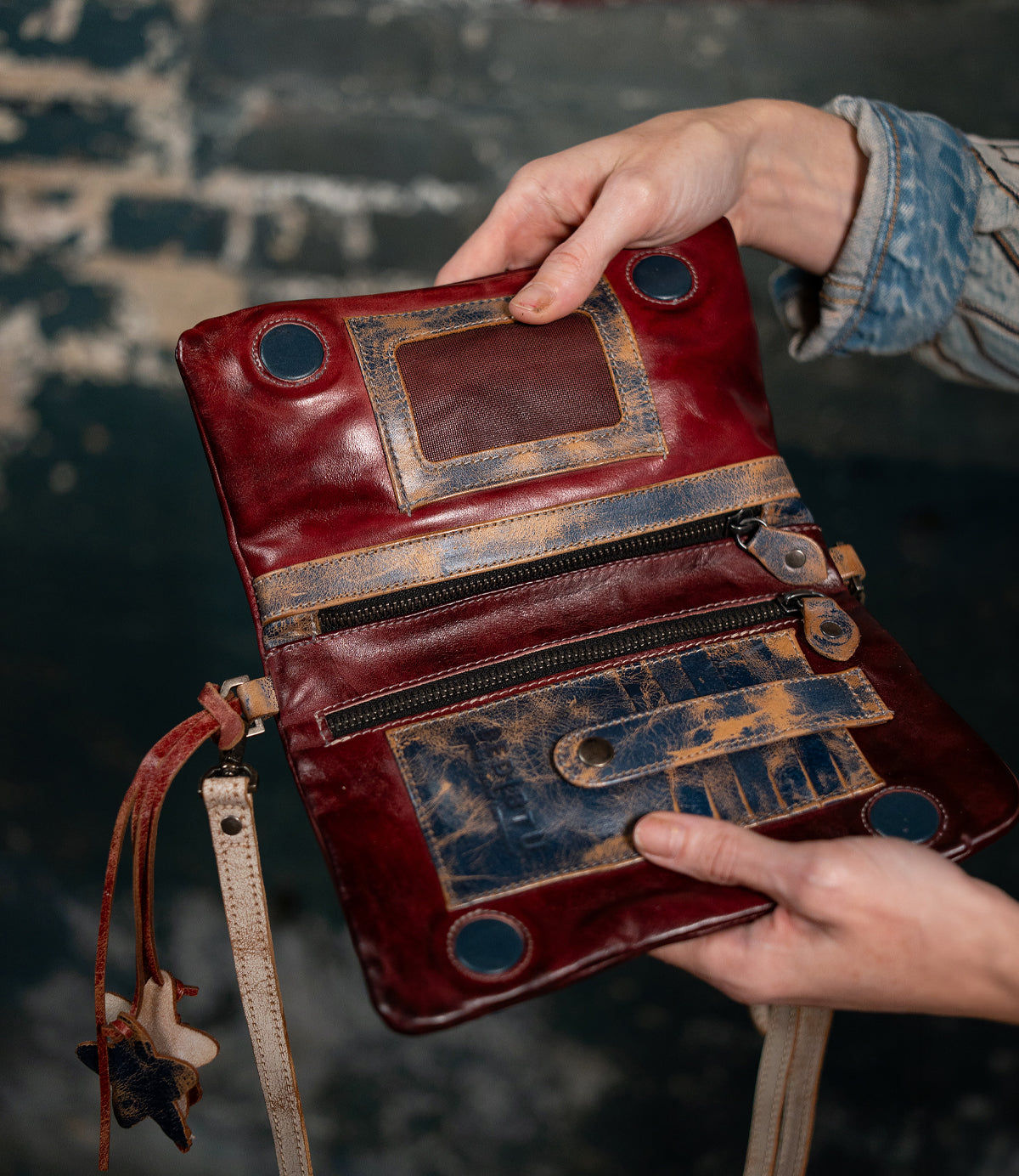 A person holding the Bed|Stü Cadence, an open vintage-style purse crafted from vegetable-tanned leather, showcasing visible zippers, roomy pockets, and distressed brown and red accents against a softly blurred dark background.