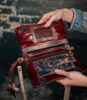 A person holding the Bed|Stü Cadence, an open vintage-style purse crafted from vegetable-tanned leather, showcasing visible zippers, roomy pockets, and distressed brown and red accents against a softly blurred dark background.