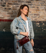 A woman wearing a denim jacket and jeans stands against a weathered brick wall, holding the Bed|Stü Cadence crossbody clutch in dark red vegetable-tanned leather.