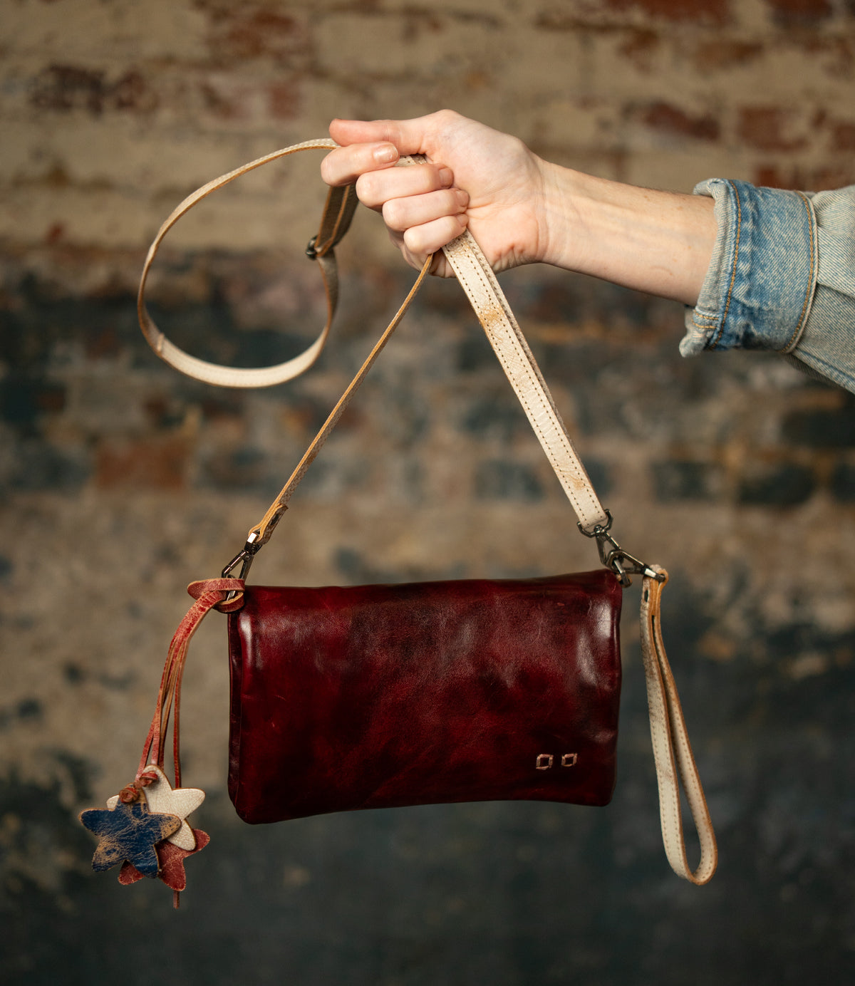 A hand holds the Bed|Stü Cadence, a compact crossbody clutch in dark red vegetable-tanned leather, featuring star-shaped leather charms and a long strap, set against a textured brick wall background.