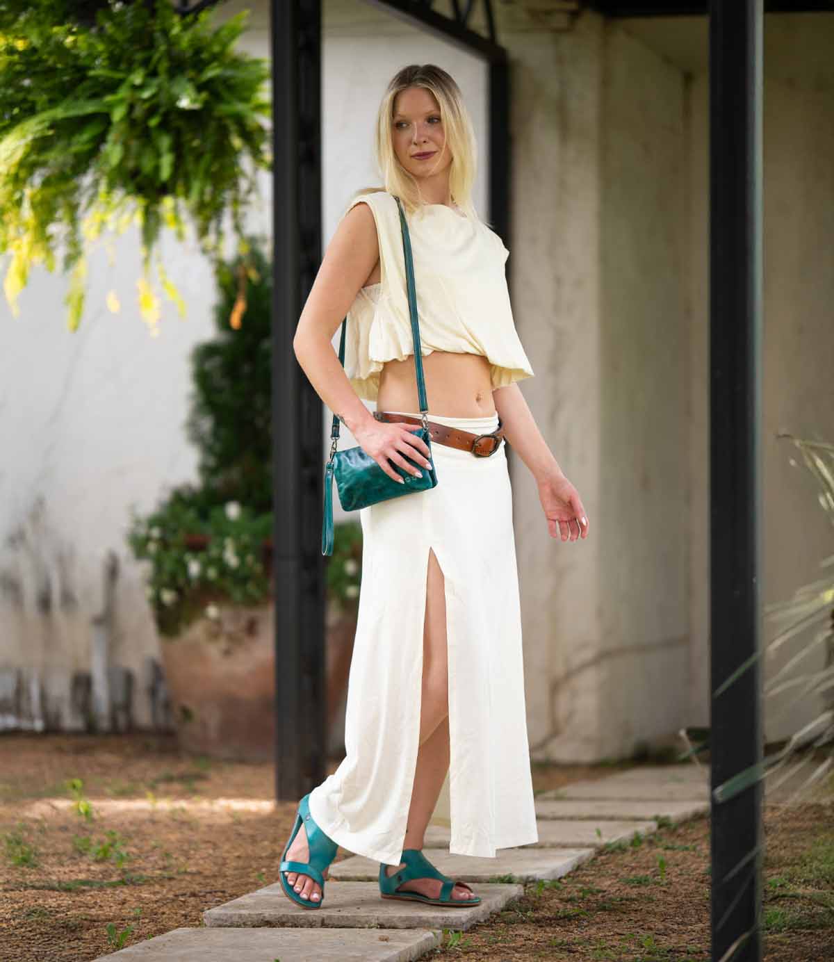 A woman stands outdoors wearing a cream sleeveless crop top, a cream skirt with a thigh-high slit, teal sandals, a brown belt, and carrying the Bed|Stü Cadence crossbody clutch crafted from vegetable-tanned leather.