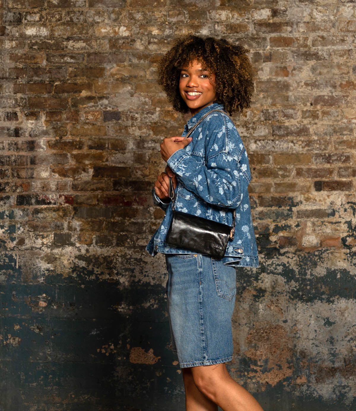 A person stands sideways against a weathered brick wall, wearing a patterned denim jacket, denim skirt, and holding the Cadence vegetable-tanned leather crossbody clutch by Bed|Stü.