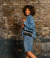 A person stands sideways against a weathered brick wall, wearing a patterned denim jacket, denim skirt, and holding the Cadence vegetable-tanned leather crossbody clutch by Bed|Stü.