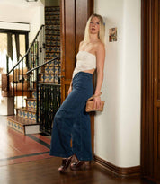 A woman stands indoors by a staircase, wearing a strapless white top, high-waisted wide-leg jeans, brown heels, and holding the Cadence crossbody clutch by Bed|Stü—crafted from vegetable-tanned leather.