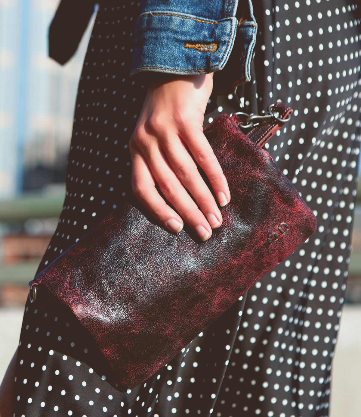 A person in a polka dot dress and denim jacket holds the Bed|Stü Cadence, a dark red crossbody clutch crafted from vegetable-tanned leather, outdoors.