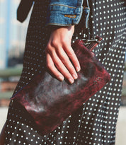 A person in a polka dot dress and denim jacket holds the Bed|Stü Cadence, a dark red crossbody clutch crafted from vegetable-tanned leather, outdoors.