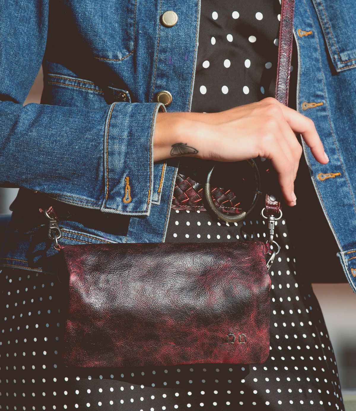 Wearing a denim jacket and polka dot dress with a woven belt at the waist, the person holds the Bed|Stü Cadence—a textured maroon crossbody clutch with a wrist strap.