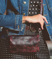 Wearing a denim jacket and polka dot dress with a woven belt at the waist, the person holds the Bed|Stü Cadence—a textured maroon crossbody clutch with a wrist strap.