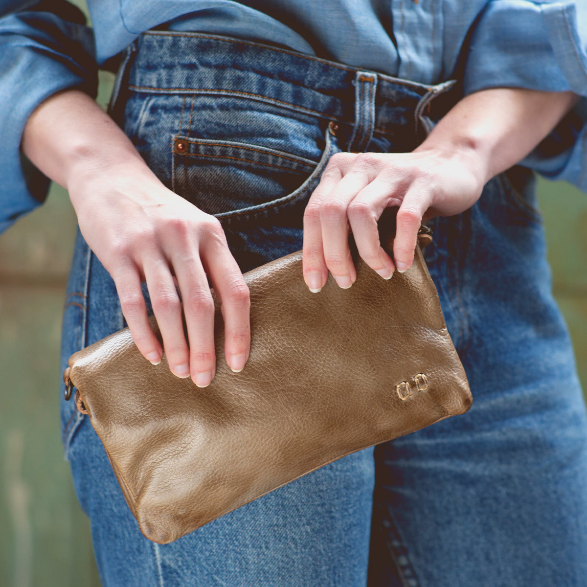 A person in a denim shirt and jeans holds the Bed|Stü Cadence brown vegetable-tanned leather crossbody clutch purse with both hands.