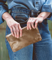 A person in a denim shirt and jeans holds the Bed|Stü Cadence brown vegetable-tanned leather crossbody clutch purse with both hands.