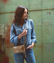 A woman in a denim shirt and jeans stands against a green, weathered metal wall, looking to the side with one hand in her pocket and carrying the Bed|Stü Cadence tan vegetable-tanned leather crossbody bag.
