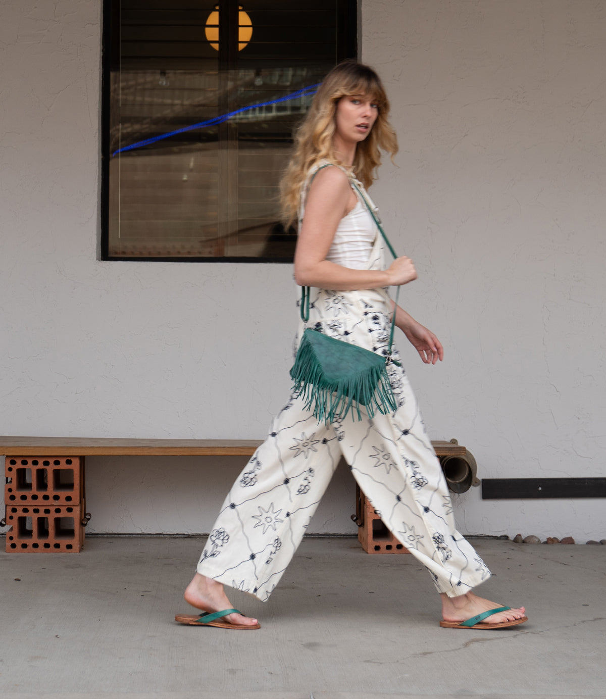 A woman in a patterned white outfit and lightweight green Calma sandals by Bed|Stü walks past a bench, carrying a green fringed bag, with a window and light fixture in the background.