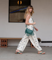 A woman in a patterned white outfit and lightweight green Calma sandals by Bed|Stü walks past a bench, carrying a green fringed bag, with a window and light fixture in the background.