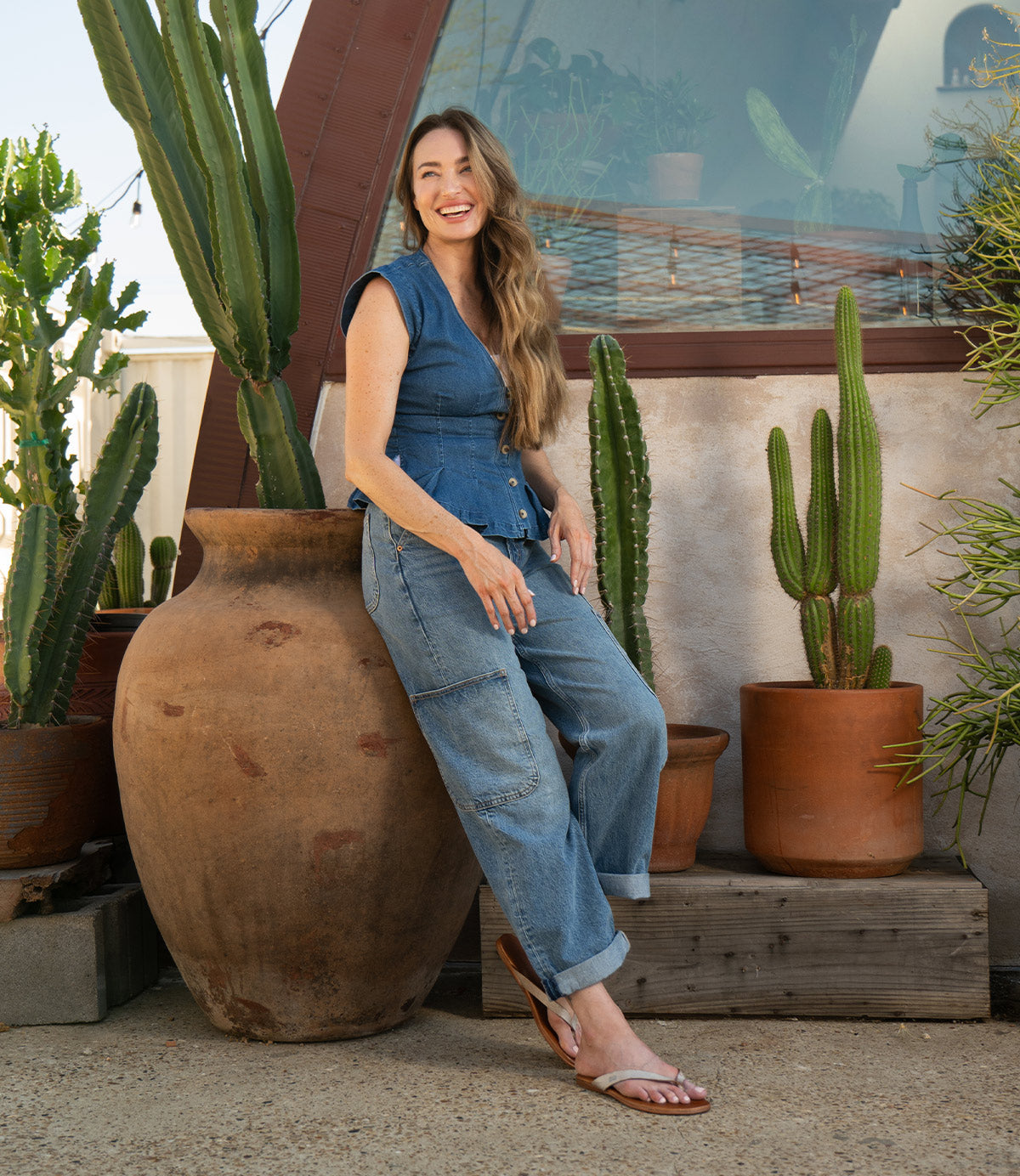 Clad in a blue denim outfit, a woman smiles beside large potted cacti under the sunny sky, her feet slipped into lightweight and comfortable Bed|Stü Calma leather sandals.
