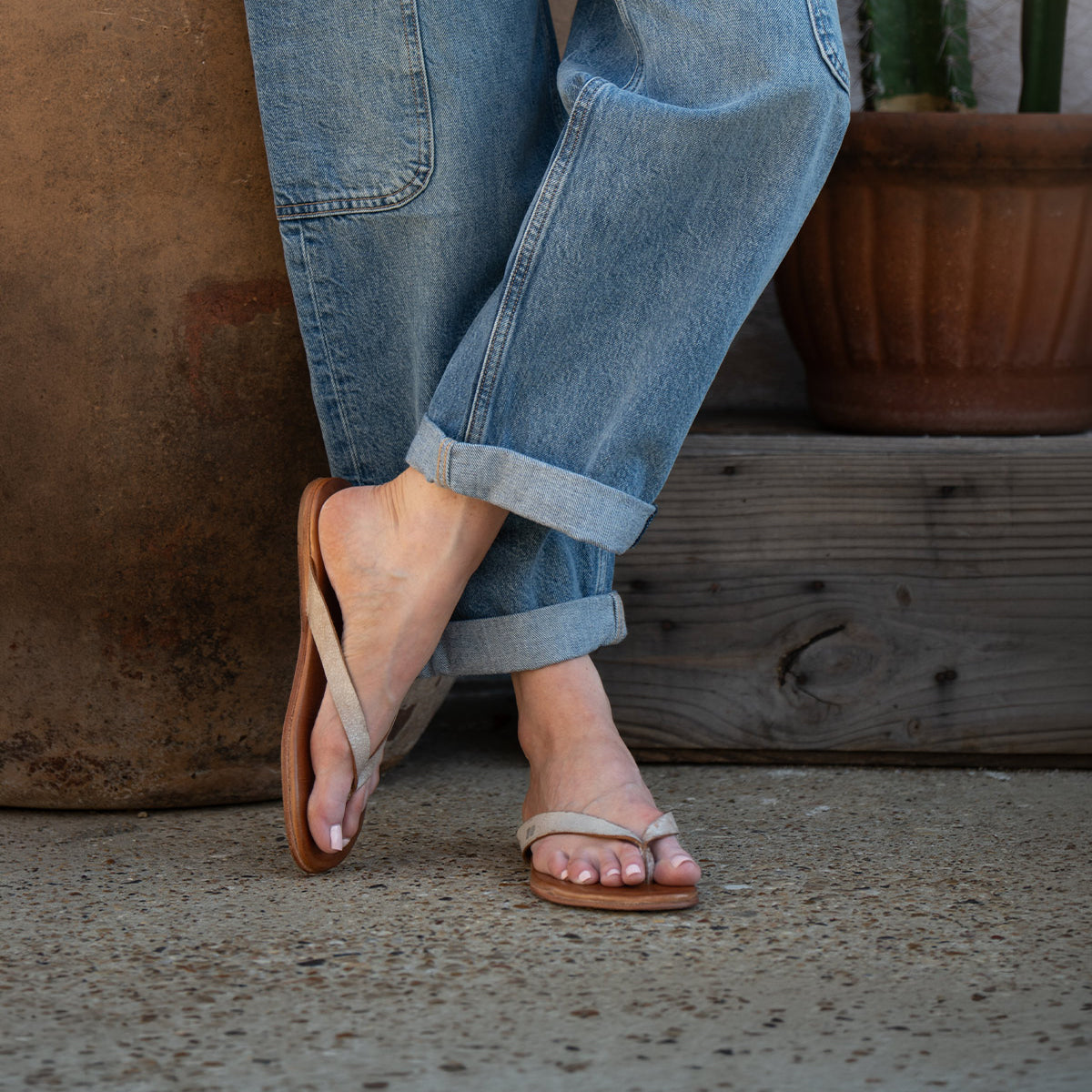 A person wearing rolled-up blue jeans and comfortable Calma beige flip-flops by Bed|Stü stands on a concrete surface next to a potted plant.