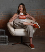 A woman sits in a white chair against a brick wall, holding the stylish orange Bed|Stü Canton 2-in-1 clutch and leather wallet, dressed in beige clothing and red patterned shoes.