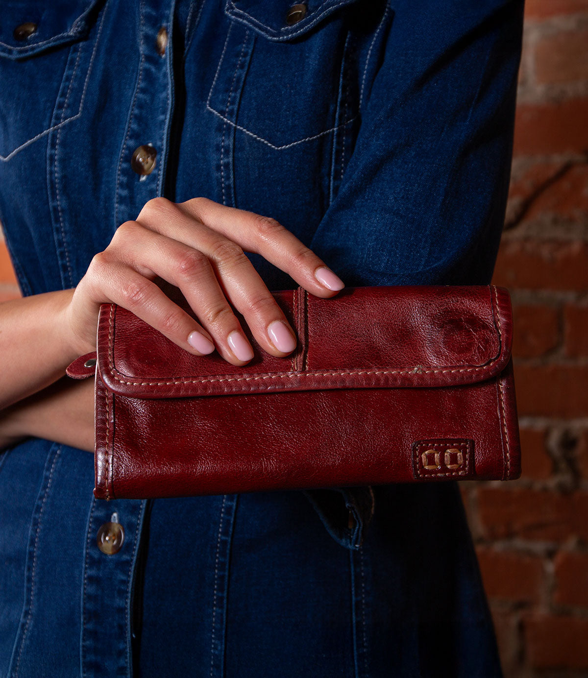 A person in a denim shirt holds the striking red Bed|Stü Canton leather wallet, featuring visible stitching and a subtle embossed logo, effortlessly showcasing this versatile 2-in-1 clutch while standing against a brick wall.