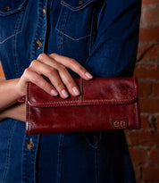 A person in a denim shirt holds the striking red Bed|Stü Canton leather wallet, featuring visible stitching and a subtle embossed logo, effortlessly showcasing this versatile 2-in-1 clutch while standing against a brick wall.