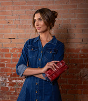 A woman in a denim dress stands in front of a brick wall, holding the chic Bed|Stü Canton 2-in-1 clutch and looking to the side with her arms crossed.