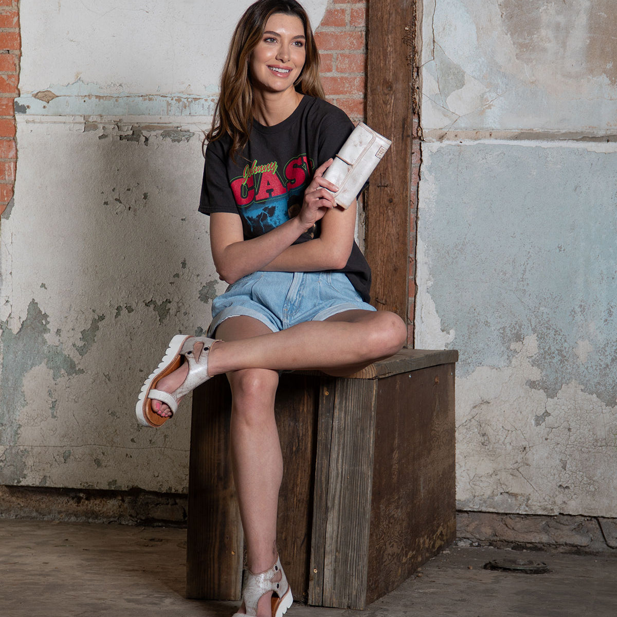 A woman sits on a wooden box in a casual outfit with a graphic t-shirt, denim shorts, and white sandals, holding the sleek Bed|Stü Canton 2-in-1 clutch against a rustic wall background.