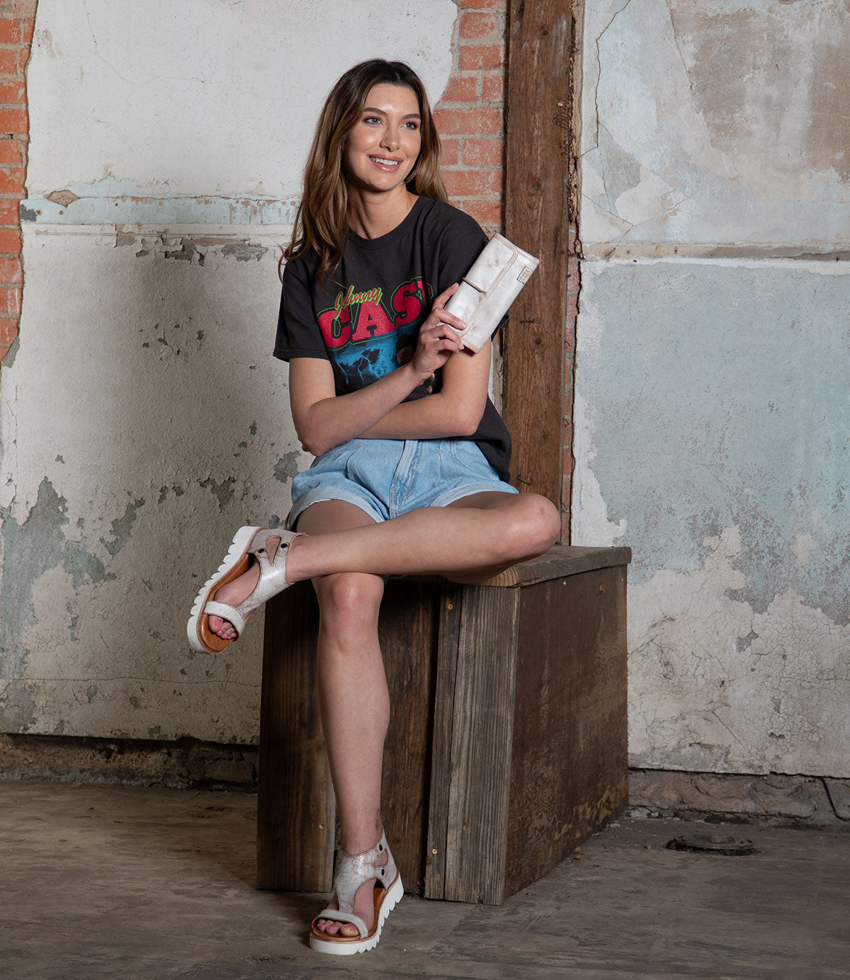 A woman sits on a wooden box in a casual outfit with a graphic t-shirt, denim shorts, and white sandals, holding the sleek Bed|Stü Canton 2-in-1 clutch against a rustic wall background.
