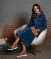 A woman in a long denim dress sits on a white chair by a brick wall, holding the Bed|Stü Canton 2-in-1 clutch and leather wallet, with flowers arranged in a metal basket on the floor beside her.
