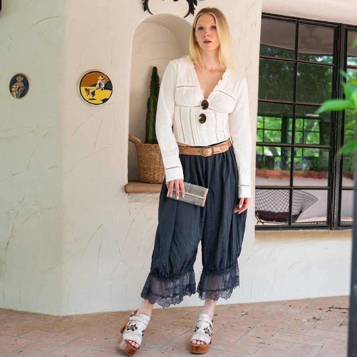A woman stands indoors by a decorated wall niche—dolphin wall decorations and a window nearby—wearing a white blouse, black lace-trimmed pants, sandals, and holding the Bed|Stü Canton chic 2-in-1 clutch.