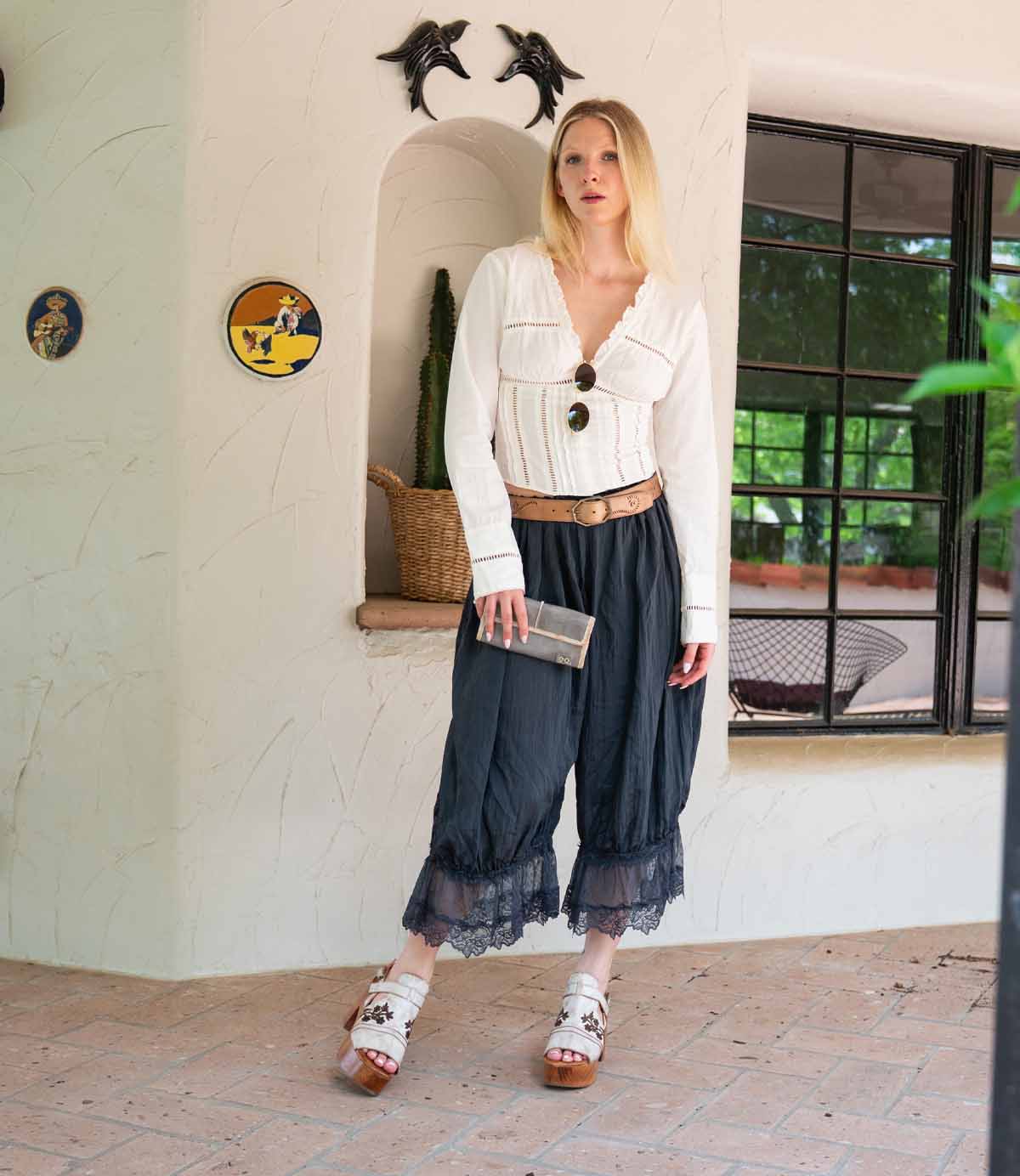 A woman stands indoors by a decorated wall niche—dolphin wall decorations and a window nearby—wearing a white blouse, black lace-trimmed pants, sandals, and holding the Bed|Stü Canton chic 2-in-1 clutch.