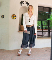 A woman stands indoors by a decorated wall niche—dolphin wall decorations and a window nearby—wearing a white blouse, black lace-trimmed pants, sandals, and holding the Bed|Stü Canton chic 2-in-1 clutch.