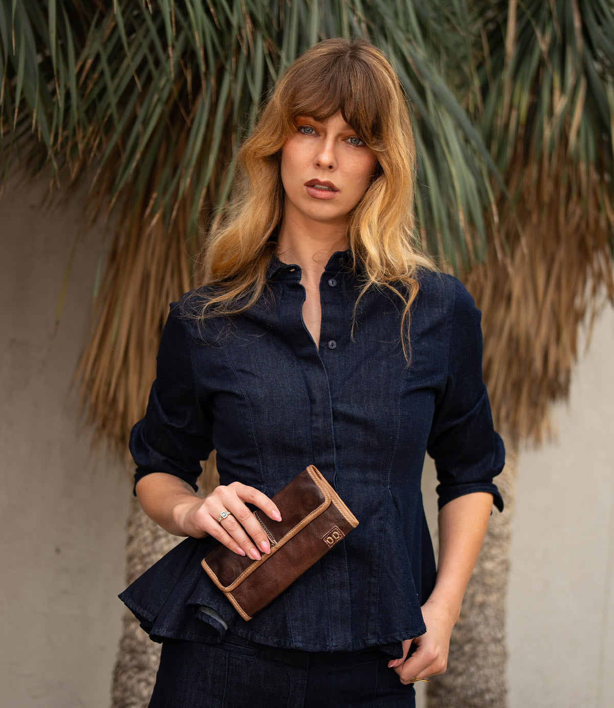 A woman with long, light brown hair wearing a dark denim shirt holds the Bed|Stü Canton leather wallet clutch while standing in front of large palm fronds.