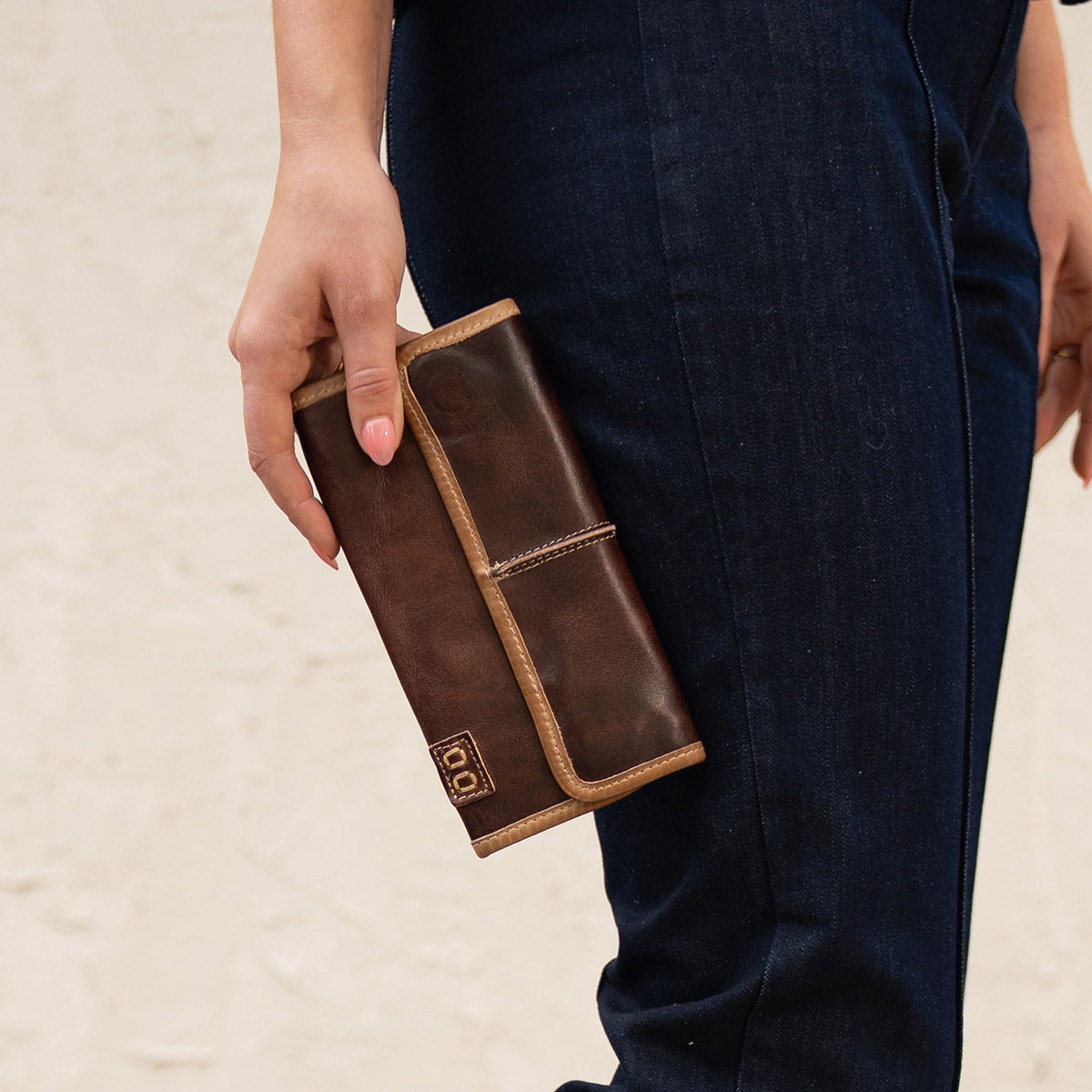 A person in dark blue jeans holds the Bed|Stü Canton, a large brown 2-in-1 leather wallet or clutch with light tan edges and a small logo on the corner.