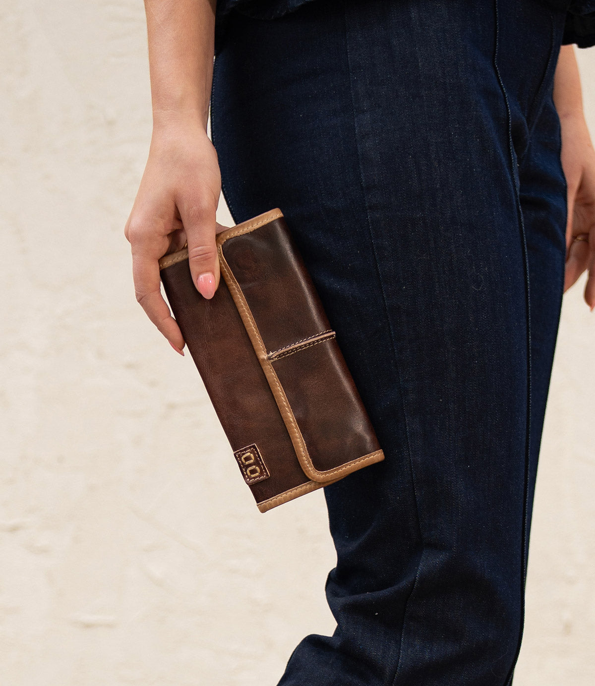 A person in dark blue jeans holds the Bed|Stü Canton, a large brown 2-in-1 leather wallet or clutch with light tan edges and a small logo on the corner.